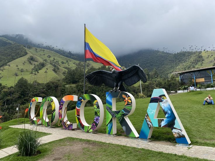 Estatua de un cóndor sobre las letras de colores de Cócora, con la bandera de Colombia y montañas del Eje Cafetero al fondo