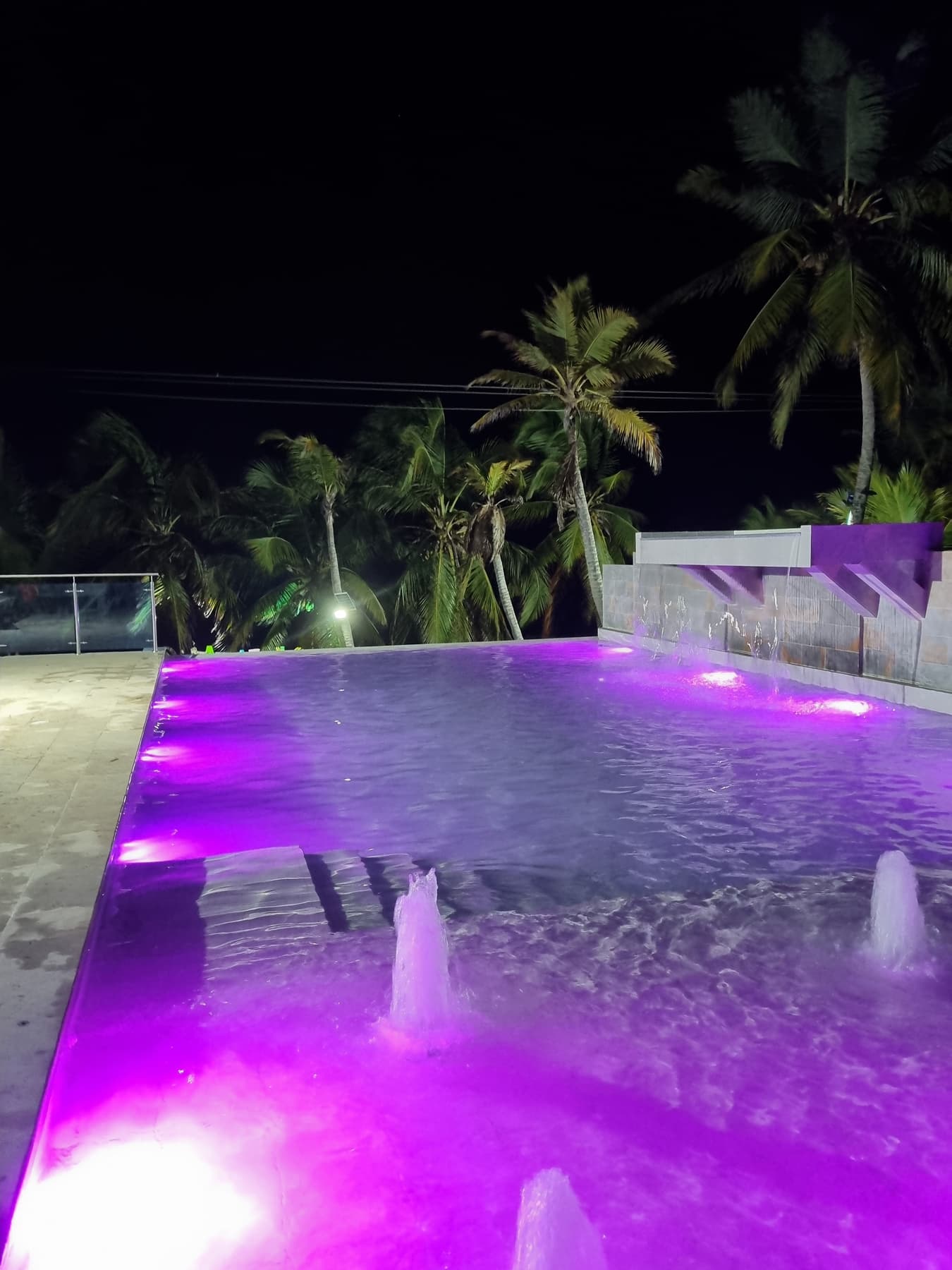 Piscina de Apartahotel Acapulco de noche, iluminada con luces moradas y chorros de agua